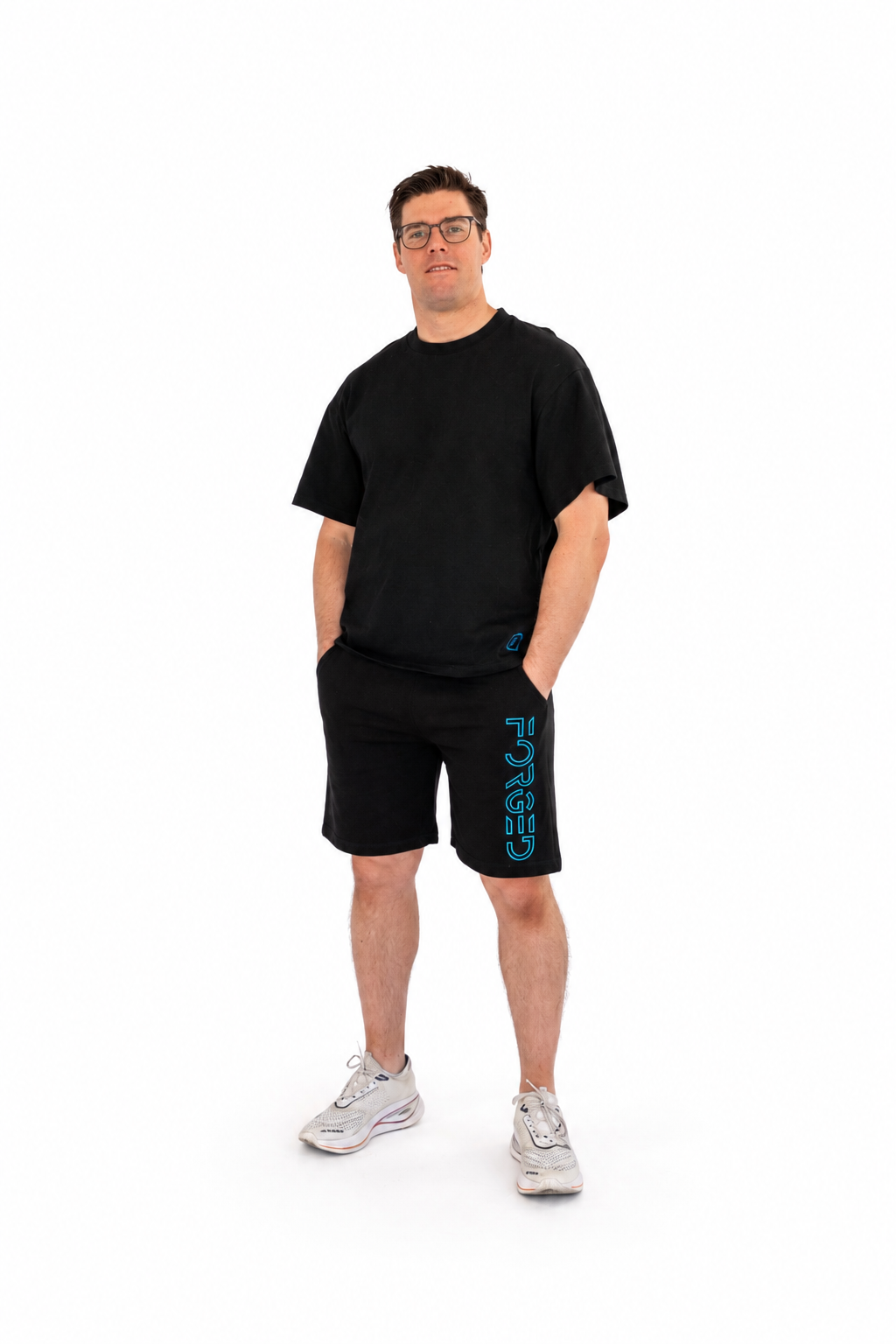 A man wearing a black crewneck t-shirt and black athletic shorts with a blue vertical 'FORGED' print on the side, paired with white sneakers, standing against a solid white background.