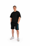 A man wearing a black crewneck t-shirt and black athletic shorts with a blue vertical 'FORGED' print on the side, paired with white sneakers, standing against a solid white background.
