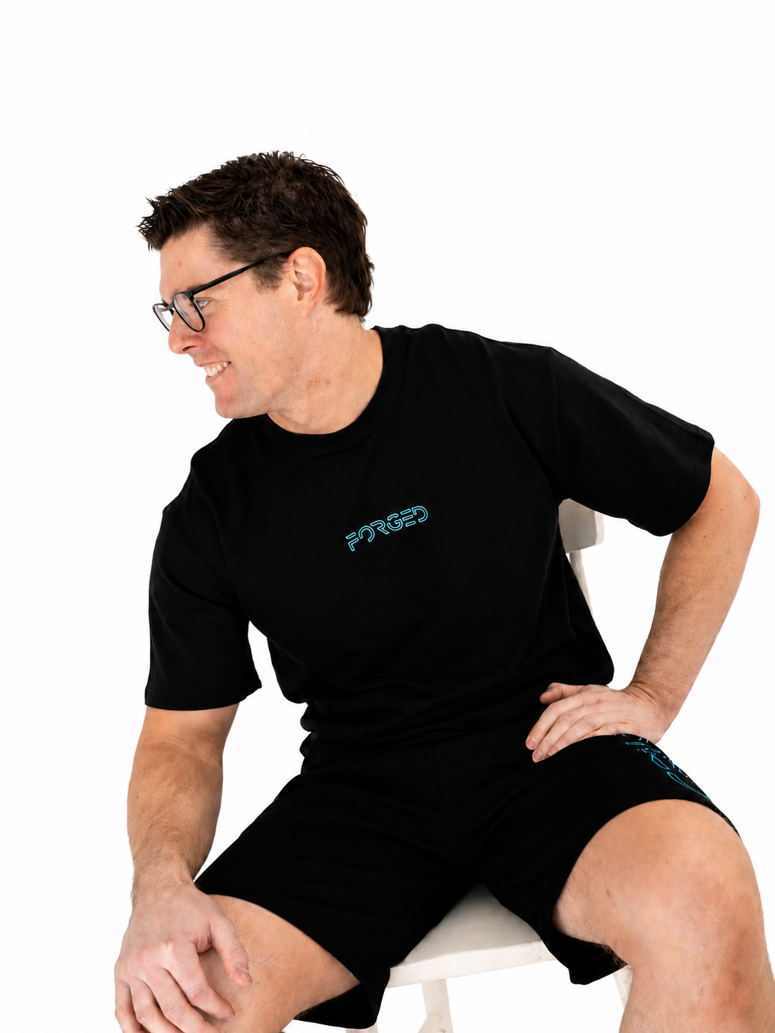 A man wearing a black short-sleeve t-shirt and matching shorts with a teal 'FORGED' logo on the chest, sitting on a white chair against a white background.
