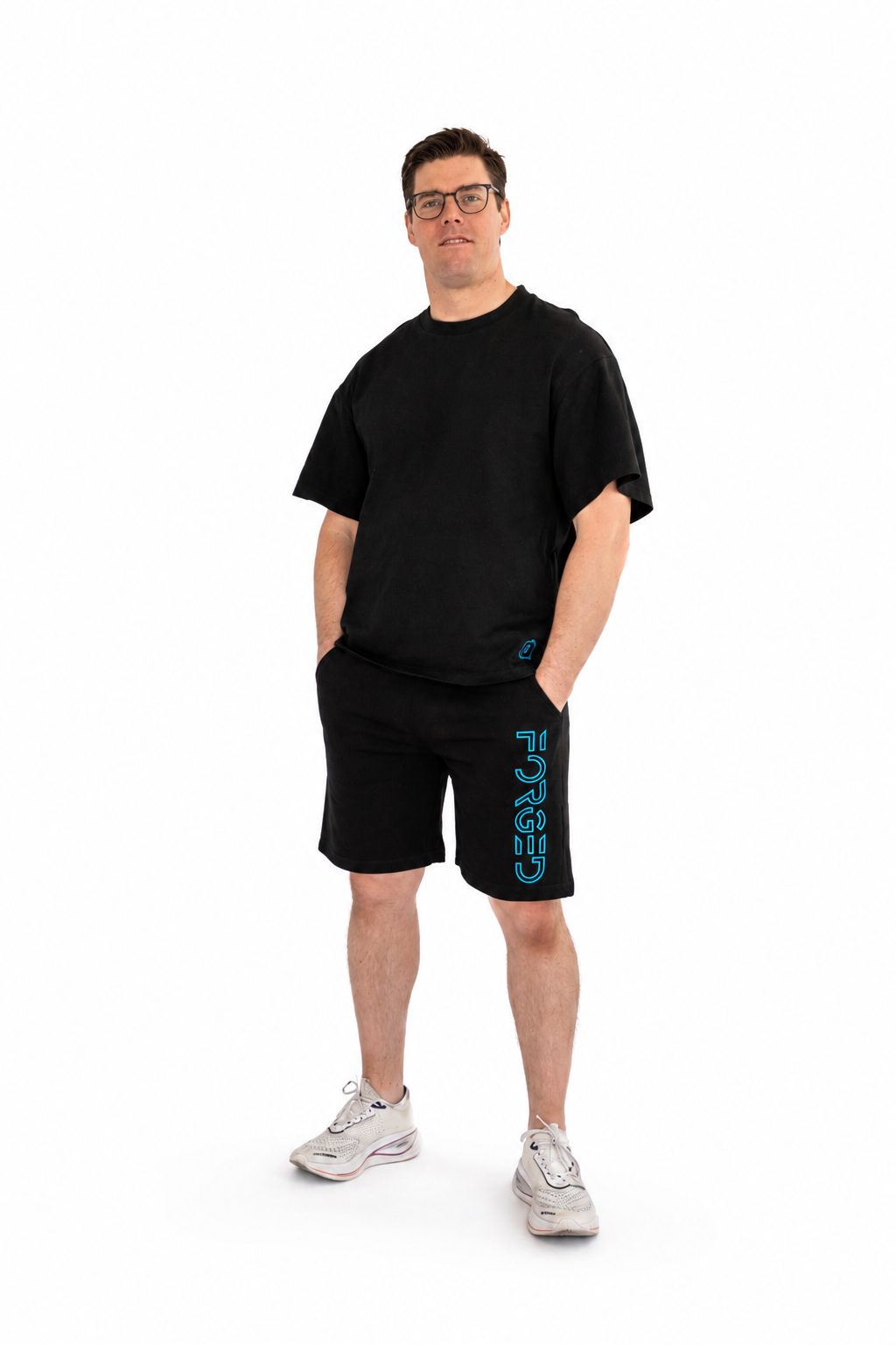 A man modeling a black short-sleeve t-shirt and matching black athletic shorts with a vertical teal 'FORGED' logo on the left leg, paired with white sneakers, against a plain white background.