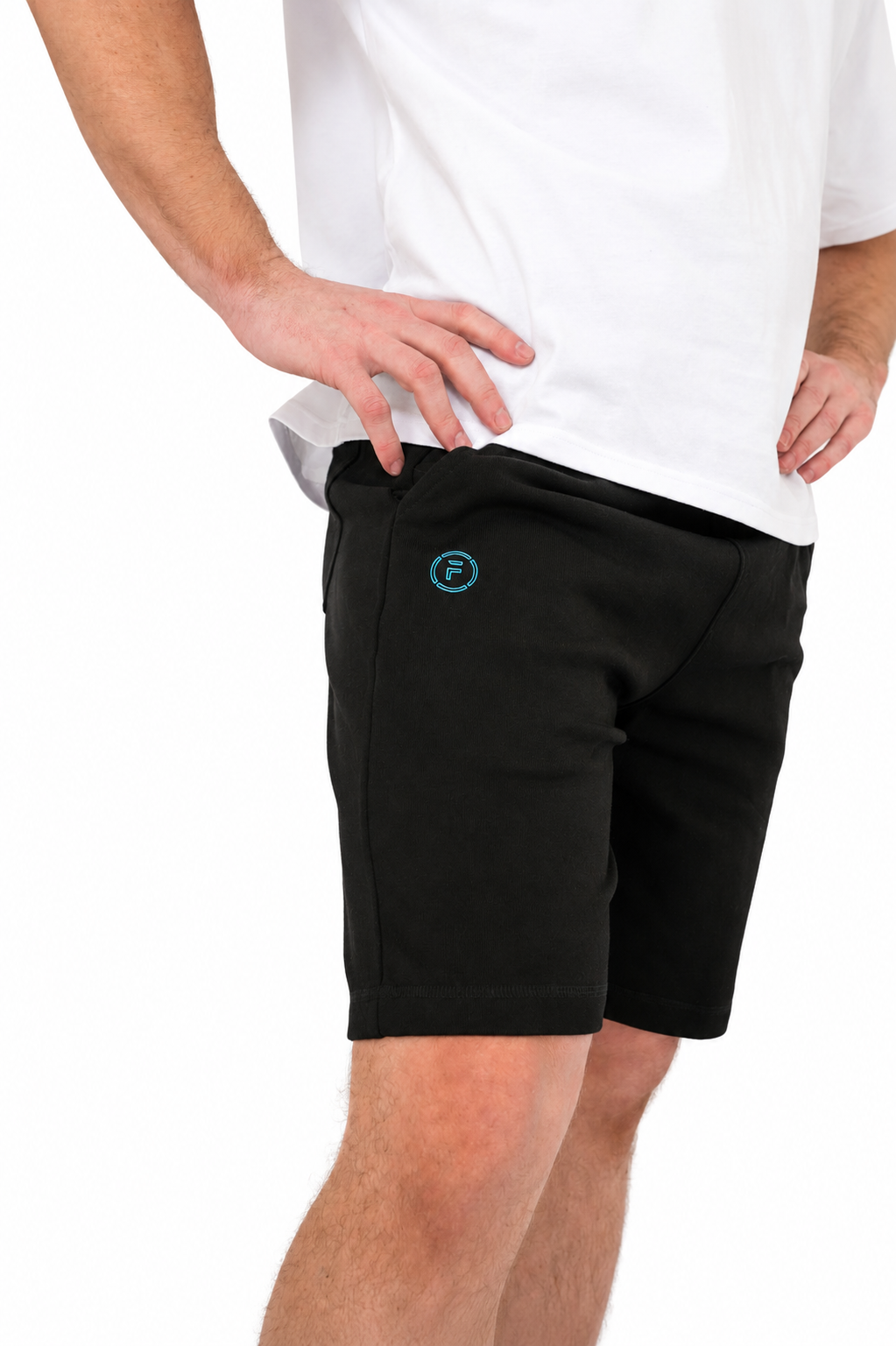 A person wearing black athletic shorts with a small teal circular logo on the left thigh, paired with a white t-shirt, shown against a plain white background.