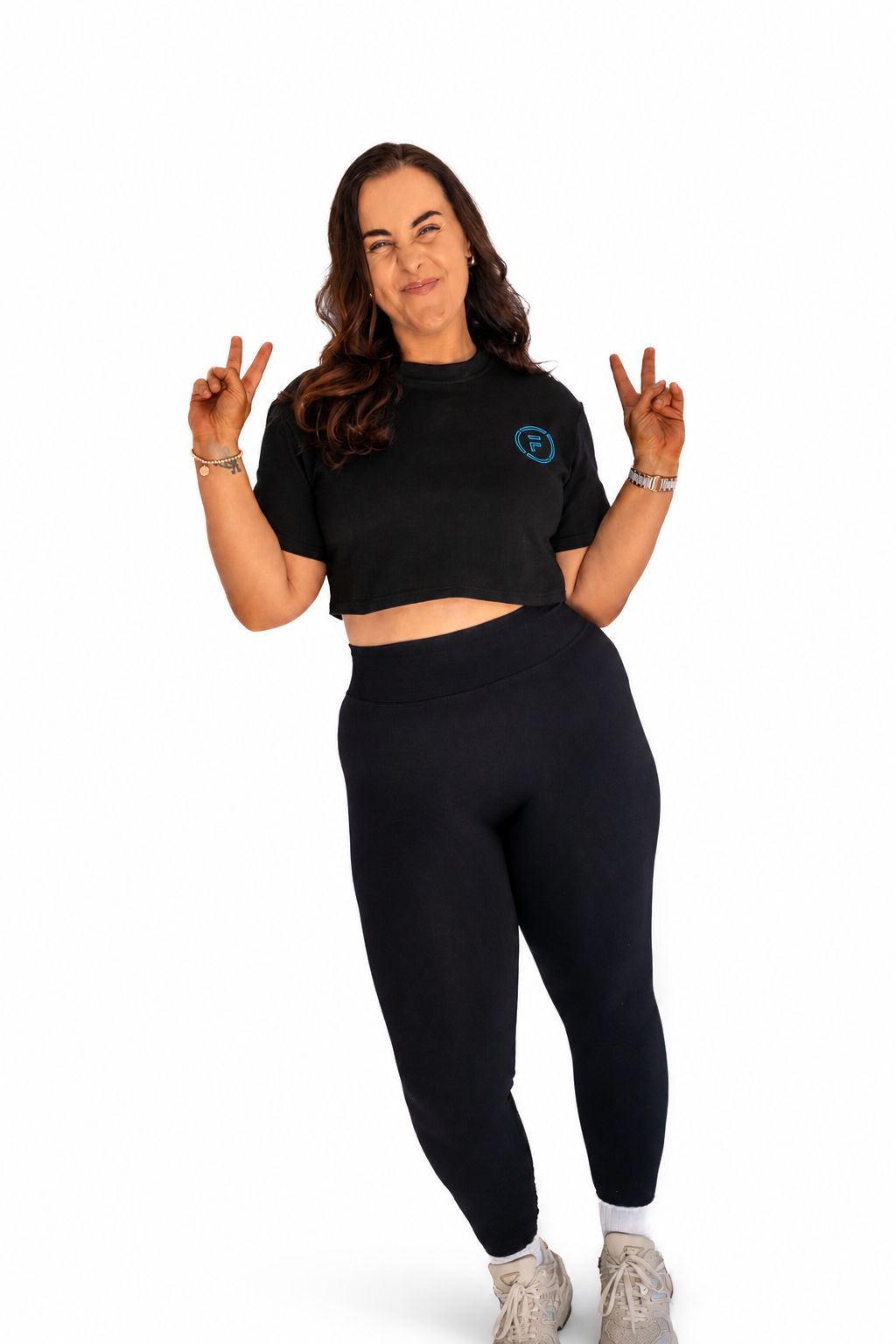 A woman modeling a black short-sleeve crop top and matching high-waisted leggings set, posing with her hands raised in a playful gesture against a solid white background.