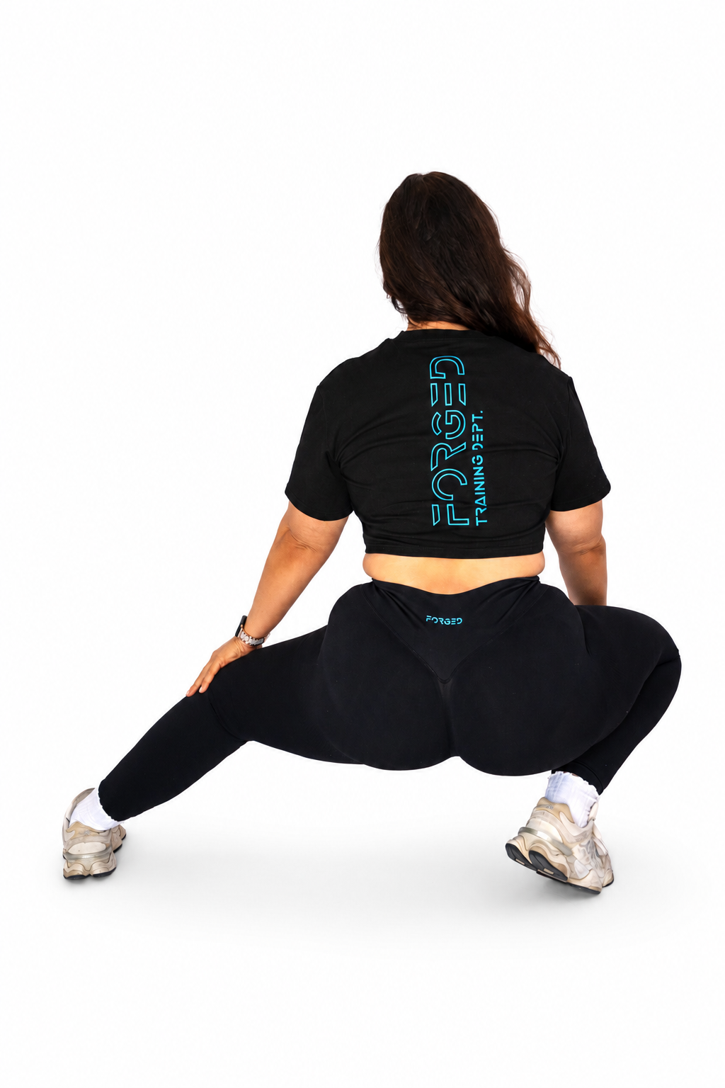 A woman from behind wearing a black crop top and black leggings, performing a deep side lunge stretch against a solid white background. The crop top features a vertical teal graphic reading 'FORGED TRAINING CLUB' on the back.