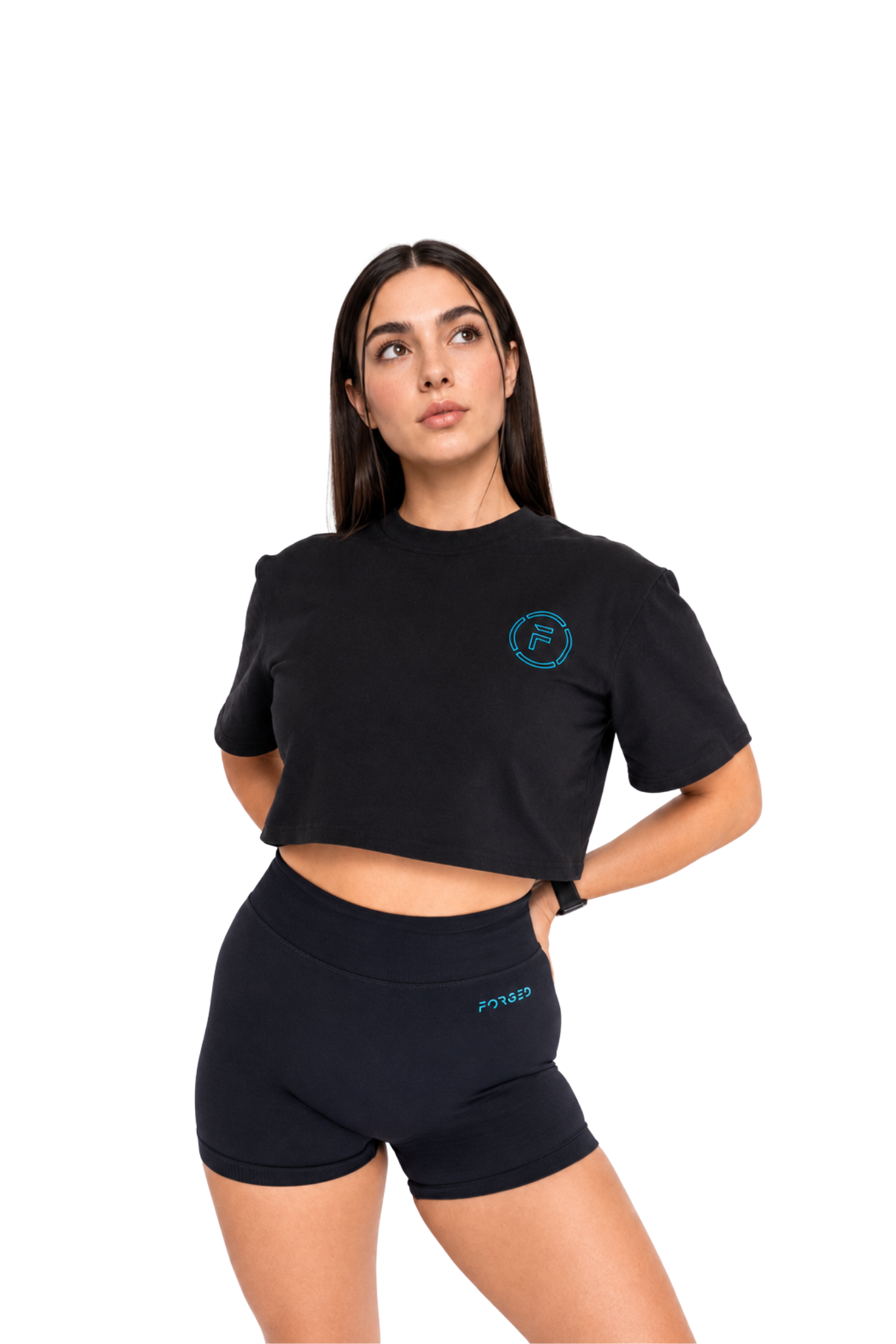 A woman modeling a black short-sleeve crop top and matching black athletic shorts against a plain white background. The crop top has a small circular teal logo on the left chest, and the shorts have a small teal wordmark on the left hip.