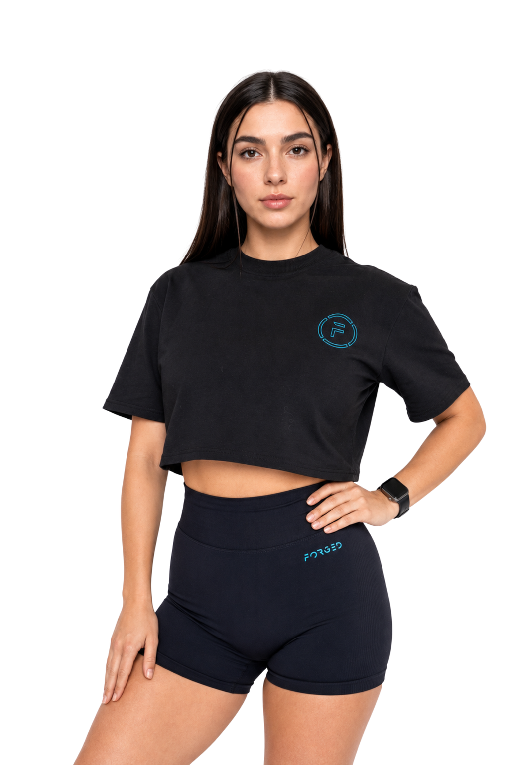 A woman modeling a black short-sleeve crop top and matching black high-waisted shorts against a plain white background. The crop top features a small teal circular logo on the left chest, and the shorts feature a small teal logo on the right hip.