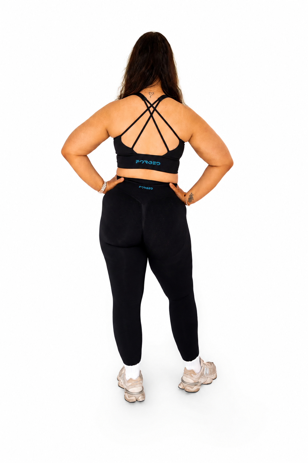 A woman is shown from the back wearing a black sports bra with crisscross straps and matching black leggings, standing against a solid white background.