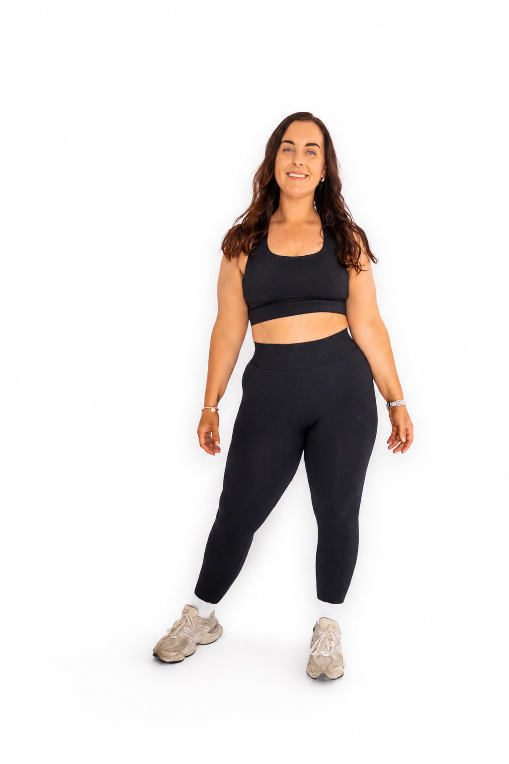 A woman modeling a matching black activewear set consisting of a sports bra and high-waisted leggings, paired with beige sneakers, against a solid white background.