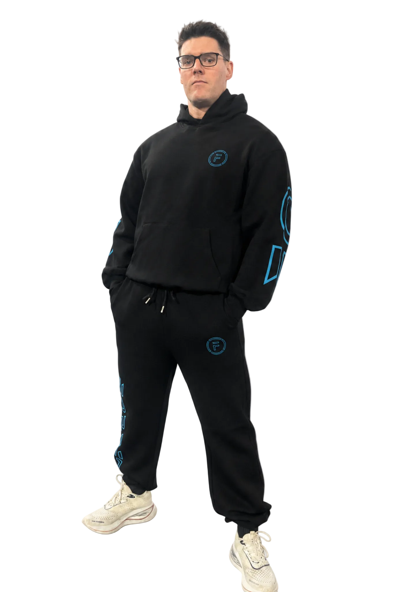 A man modeling a black two-piece tracksuit with bright blue graphic accents, consisting of a pullover hoodie and matching sweatpants, paired with beige sneakers against a light gray background.