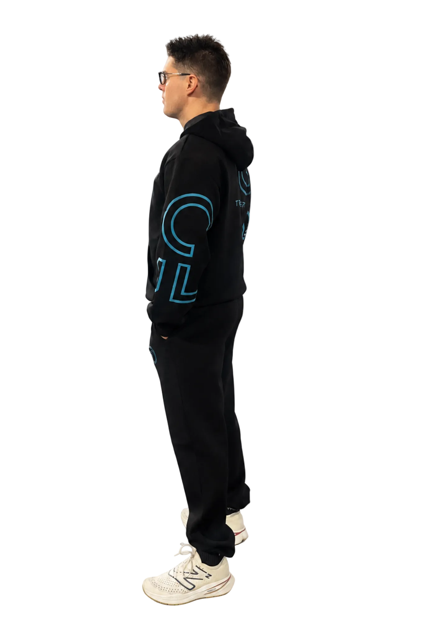 A man wearing a black hoodie and matching sweatpants with a large blue graphic on the sleeve, standing in profile against a white studio background.