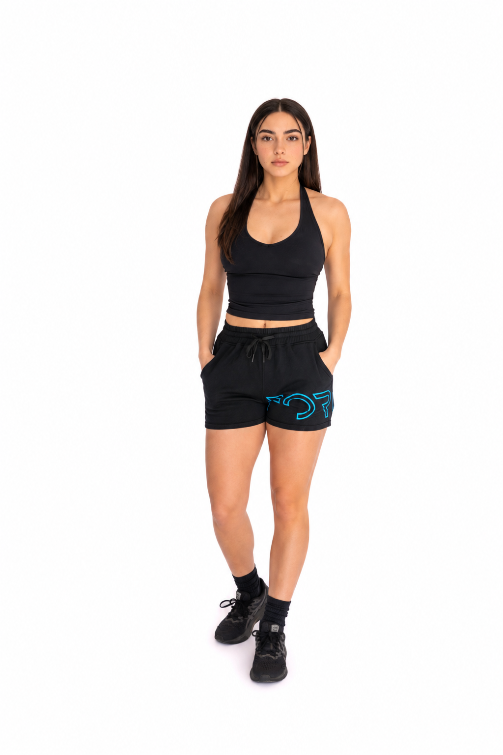 A woman modeling a black athletic set consisting of a halter crop top and matching shorts with a blue graphic logo on the left leg, paired with black sneakers, against a plain white background.