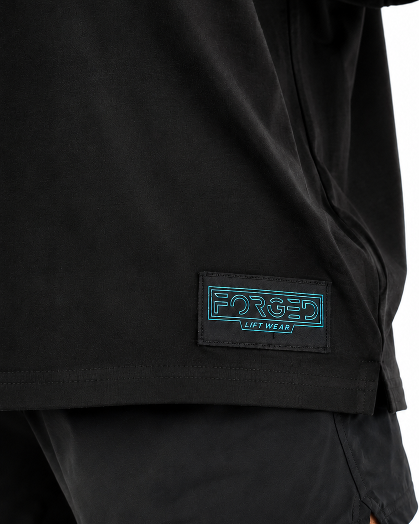 Close-up of a person wearing black shorts with a black rectangular patch on the left leg featuring the brand name 'FORGED' in blue outlined lettering and 'LIVE ON LAND' below, against a plain white background.