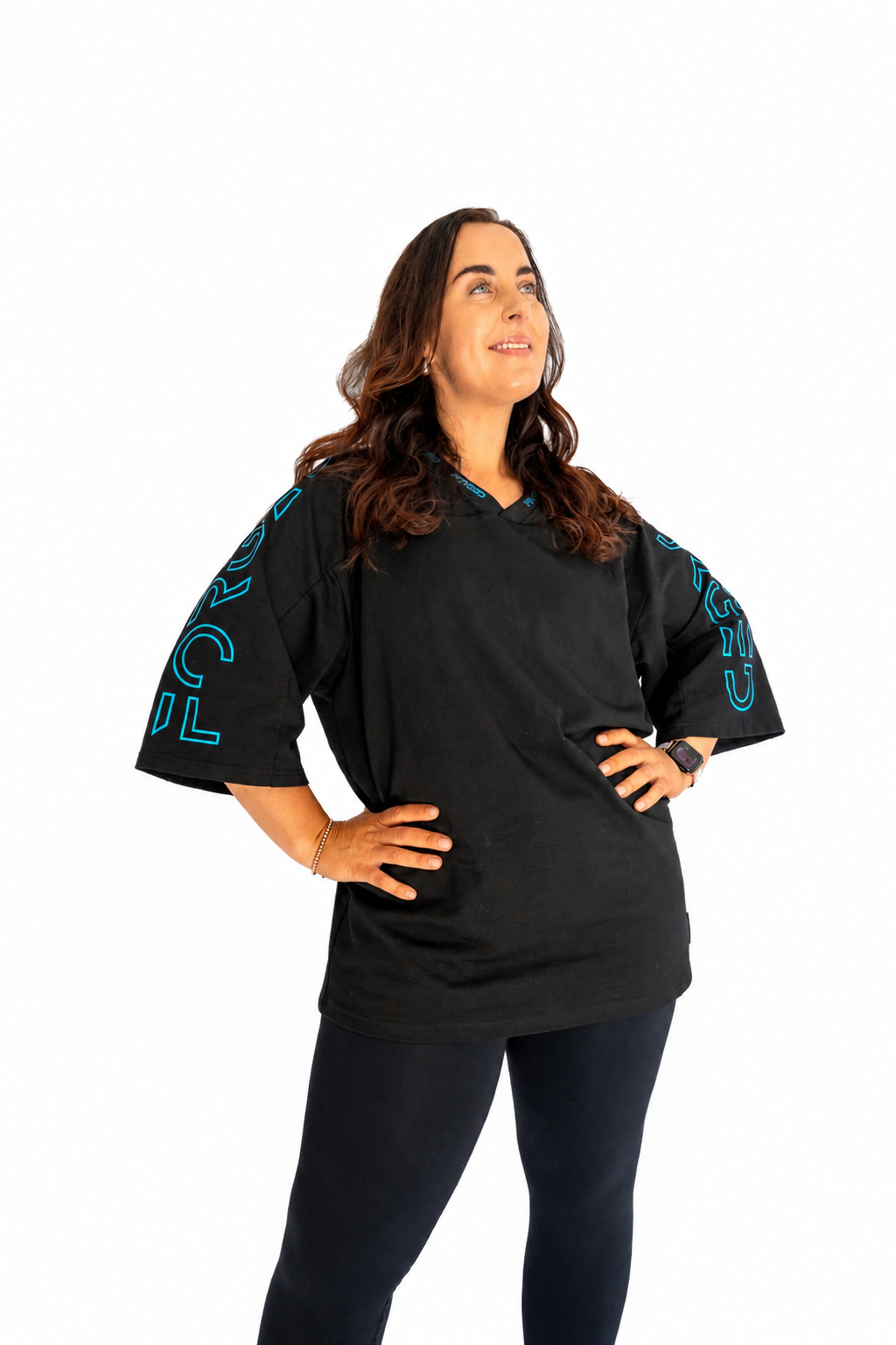 A woman modeling a black short-sleeve t-shirt with bright teal graphic lettering on the sleeves, paired with dark pants, against a solid white background.