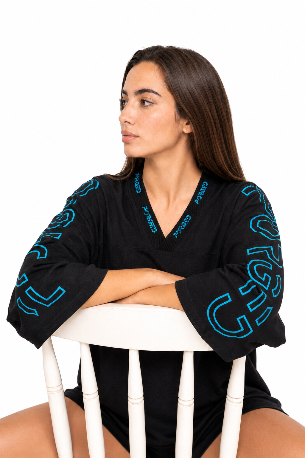 A woman with long brown hair sits on a white wooden chair, wearing a black V-neck long-sleeve top with bright blue graphic lettering on the sleeves and collar. The background is solid white.