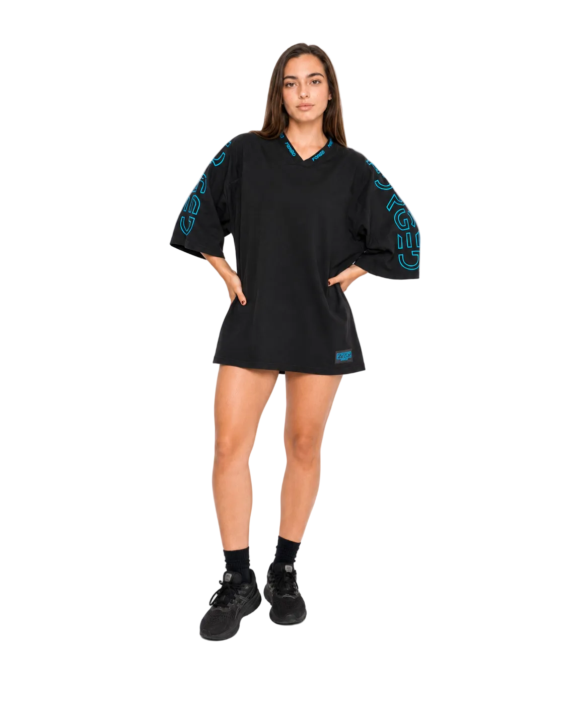 A woman modeling an oversized black short-sleeve V-neck dress with teal and pink graphic lettering on the sleeves, paired with black socks and sneakers, against a plain white studio background.