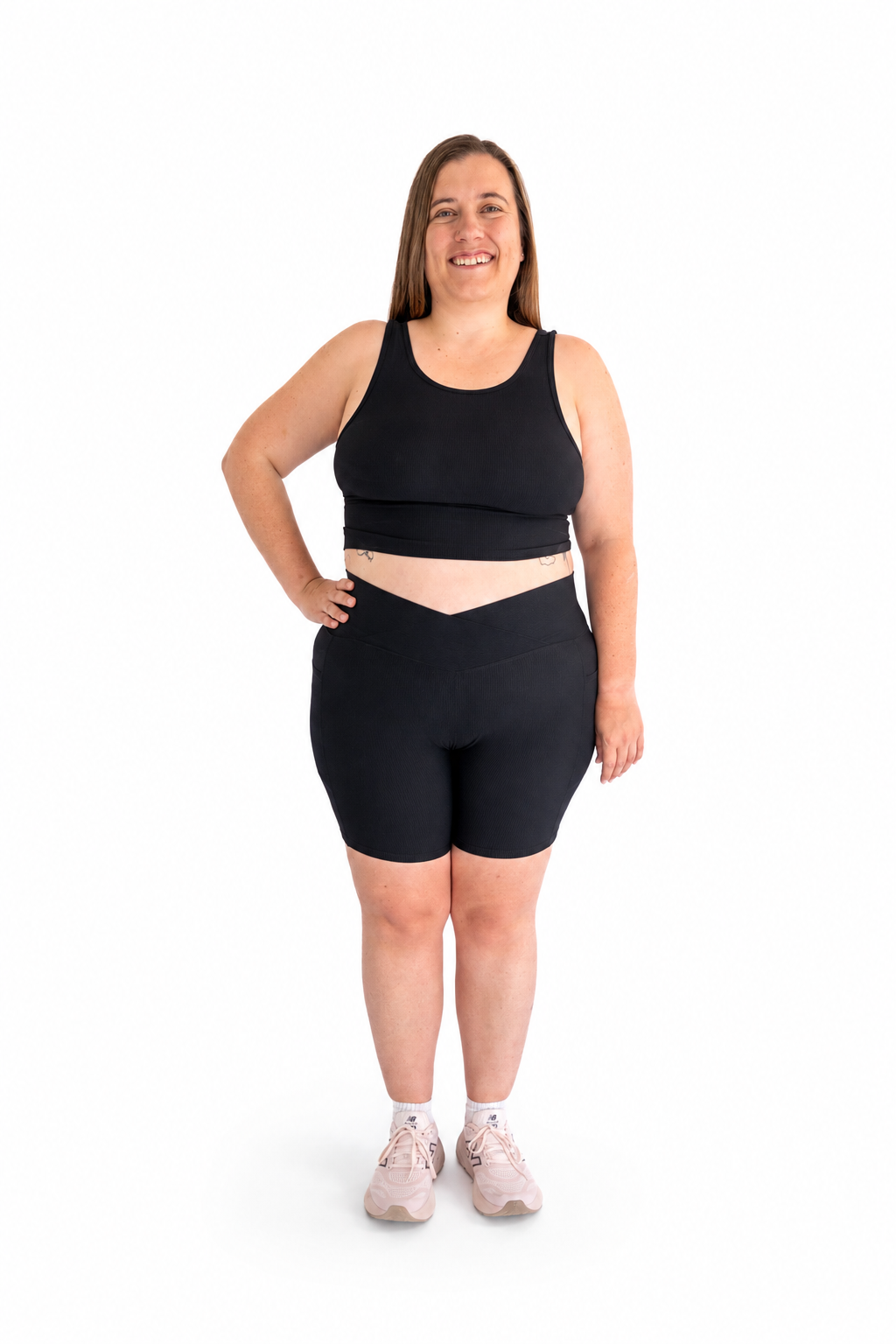 A woman modeling a matching black activewear set consisting of a cropped tank top and high-waisted biker shorts, paired with light pink sneakers, against a solid white background.