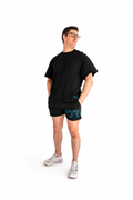 A man wearing a black oversized t-shirt, black athletic shorts with a teal graphic, and white sneakers, standing against a solid white background.
