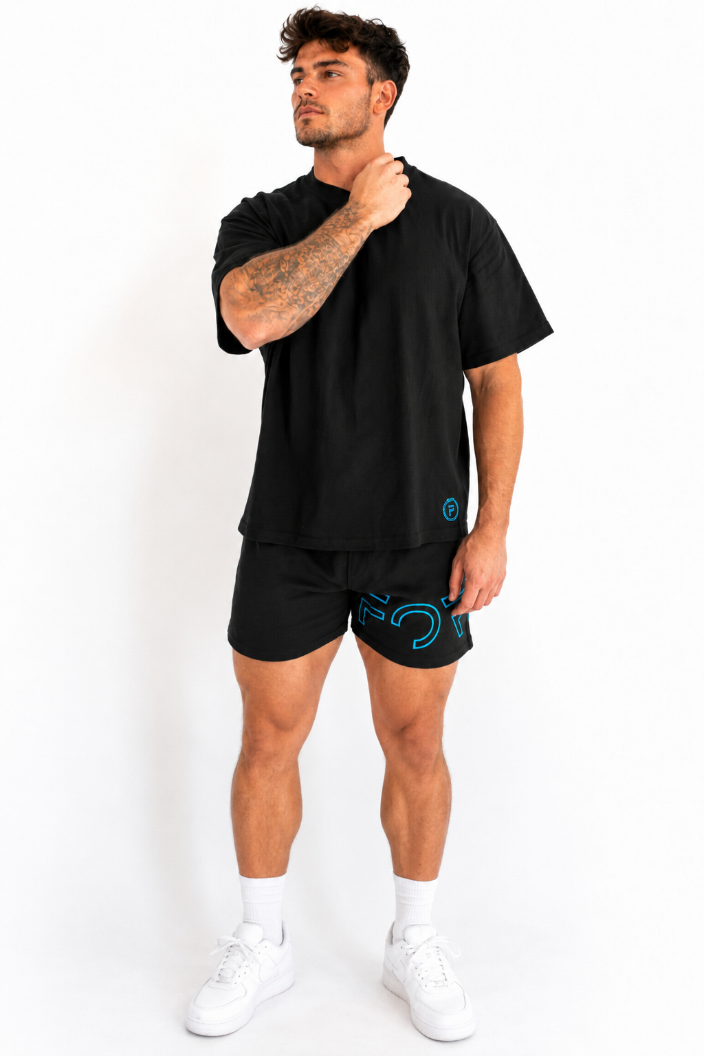 A man modeling a black short-sleeve t-shirt and matching black athletic shorts with a bright blue graphic logo on the shorts and a small blue logo on the shirt, paired with white socks and white sneakers, against a plain white background.