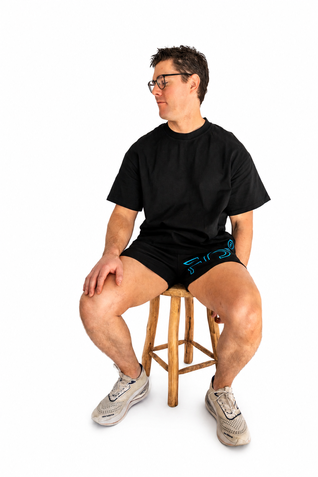 A man wearing a black t-shirt, black shorts with a blue graphic, and gray sneakers sits on a wooden stool against a solid white background.