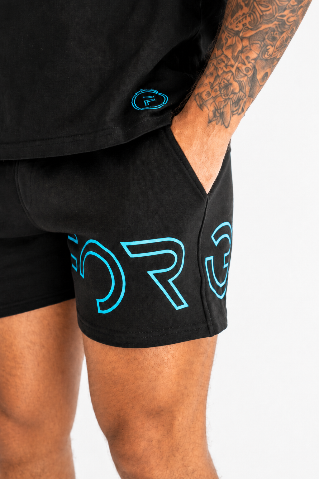 Close-up of a person wearing black athletic shorts with large bright blue outlined lettering on the side and a matching blue circular logo near the hem, against a light gray background.