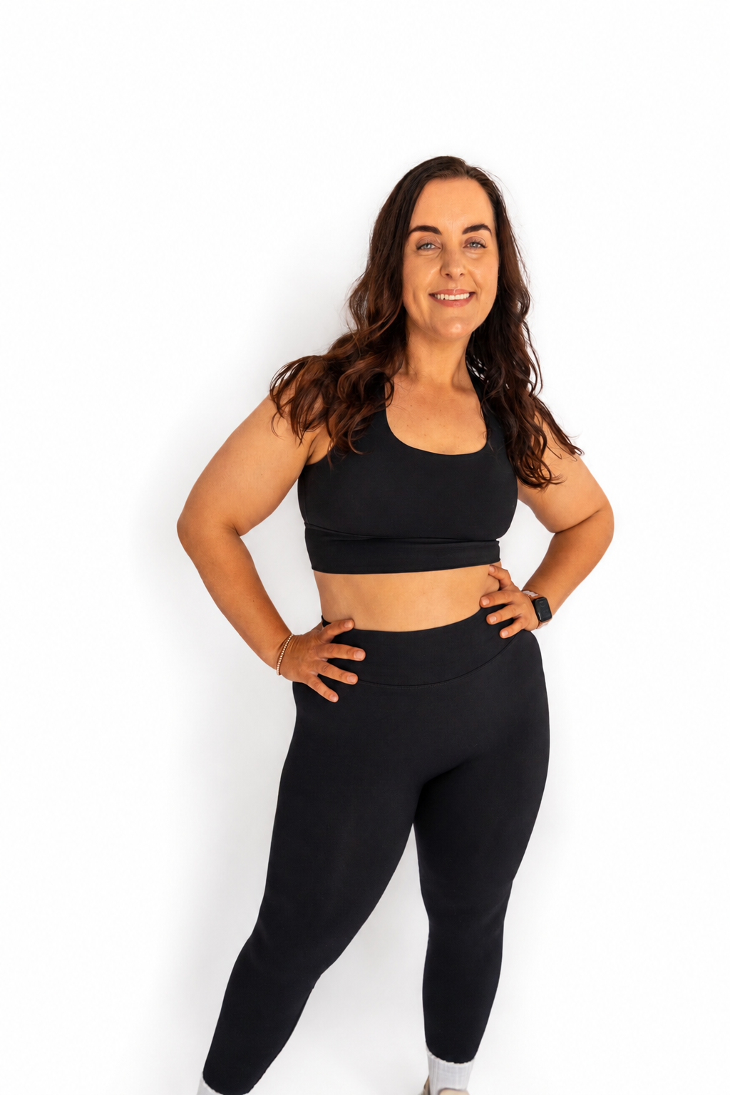 A woman modeling a matching black activewear set consisting of a sports bra and high-waisted leggings, standing against a plain white background.