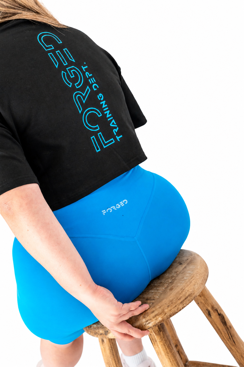 A person seated on a wooden stool wearing a black cropped T-shirt with large turquoise text and bright blue leggings. The photo is cropped from the back/side against a clean white background, with the person's hand resting on the stool.