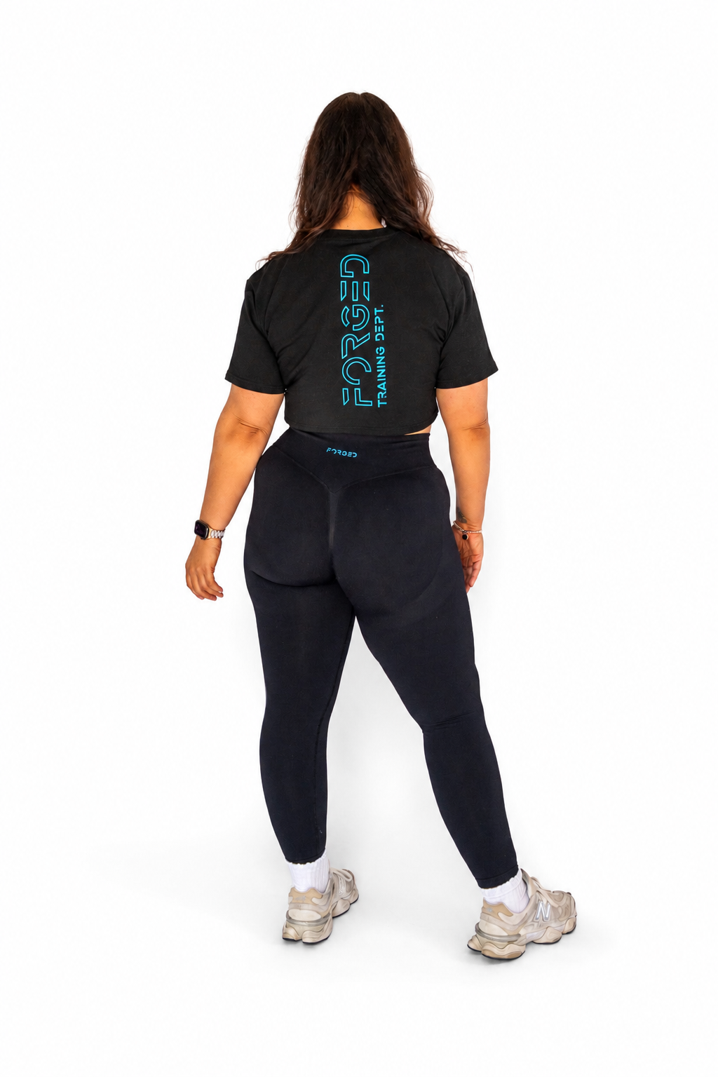 A woman is shown from behind wearing a black short-sleeve crop top with a vertical teal 'FORGED' logo and matching black leggings, standing against a plain white background.