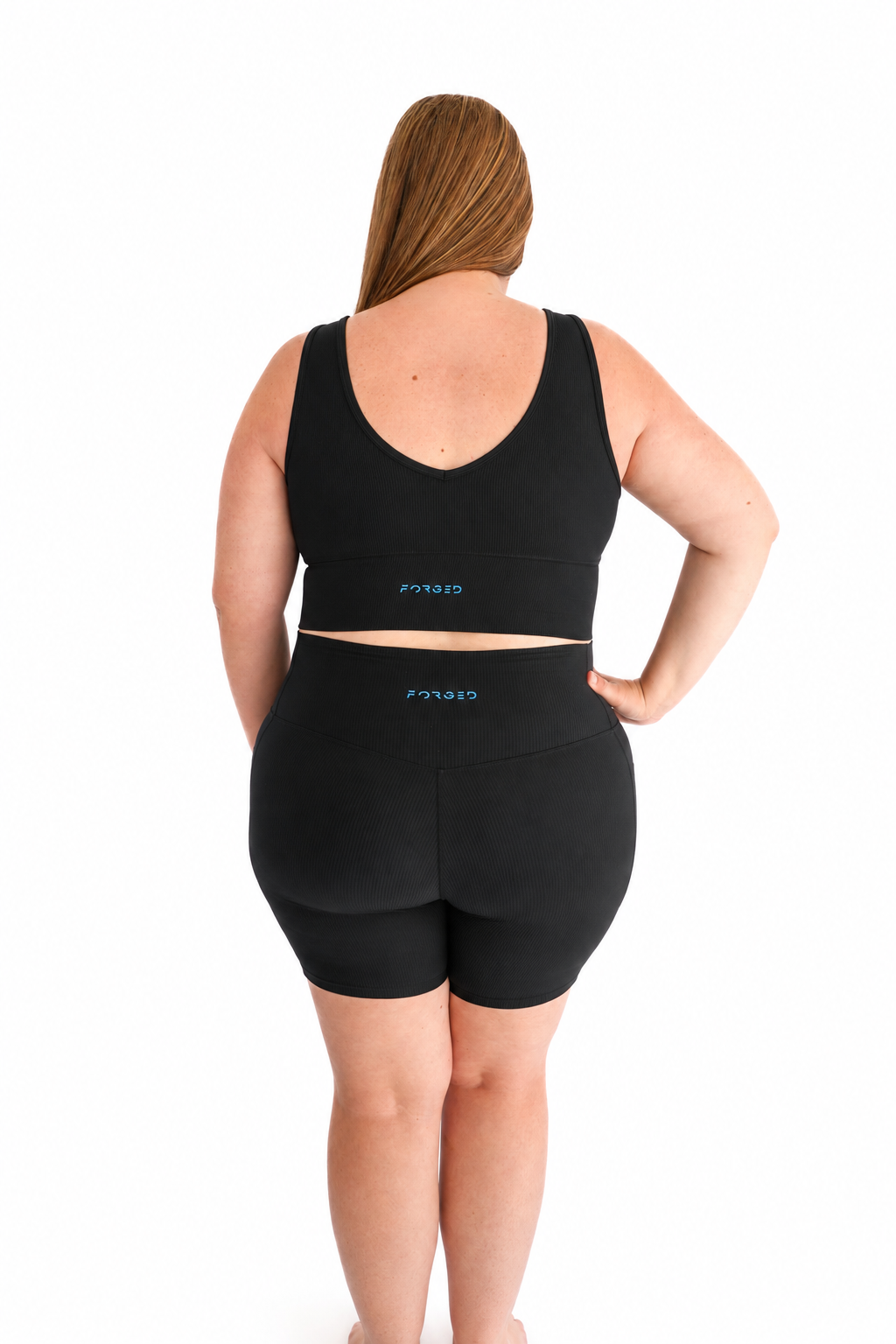 A woman shown from the back wearing a matching black activewear set consisting of a sports bra and high-waisted biker shorts, with a small blue logo on the waistband and back of the bra. She is standing against a solid white background.