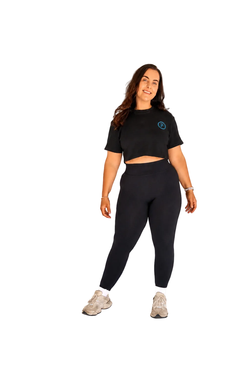 A woman modeling a black short-sleeve crop top and matching black leggings set, paired with beige sneakers, against a solid white background.