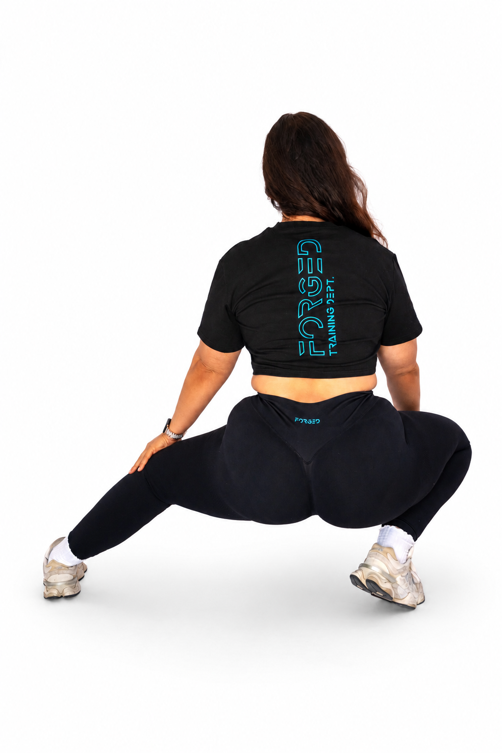 A woman from behind wearing a black crop top and leggings with a teal vertical 'FORGED' logo, performing a deep side lunge on a white studio background.